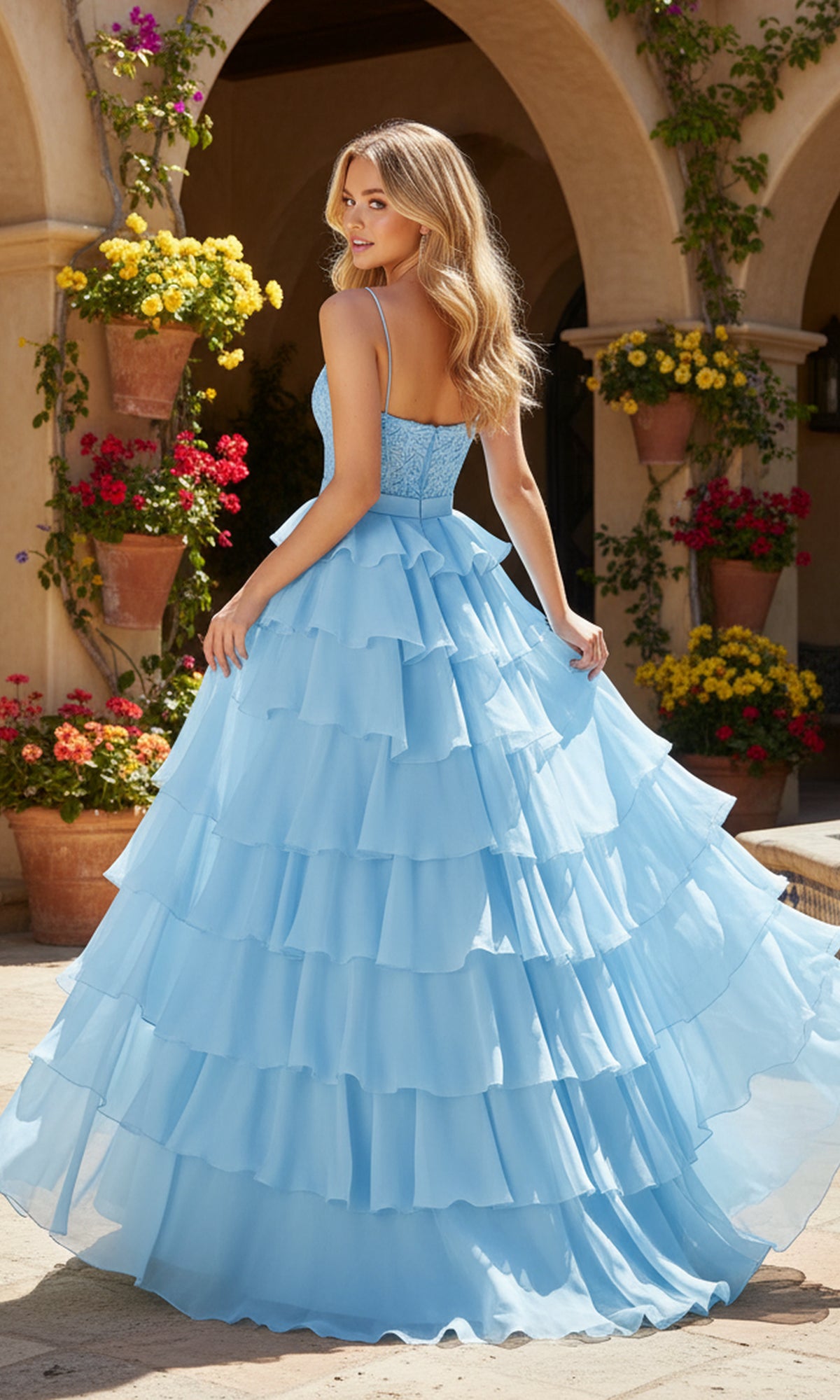 A woman wears the Long Prom Dress: PromGirl Exclusive 250202, a light blue ruffled ball gown with a lace corset, standing on a stone patio amid flowering potted plants and an arched building.